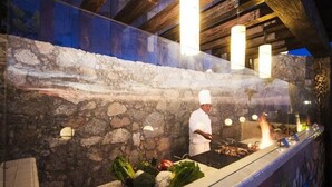 Un chef cuisinant sur un barbecue dans une cuisine extérieure dotée de murs en pierre et d’éclairages suspendus.