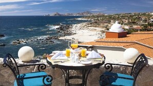 Une table avec des aliments pour le petit-déjeuner et deux chaises, donnant sur une zone côtière avec des rivages rocheux et une vue sur des montagnes lointaines.