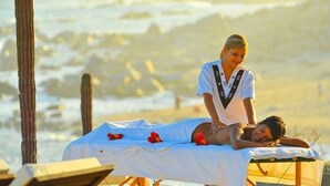 Une séance de massage sur une plage, avec une personne allongée sur une table de massage, une autre personne qui s’occupe d’elle, et en arrière-plan, des cactus et les vagues de l’océan.