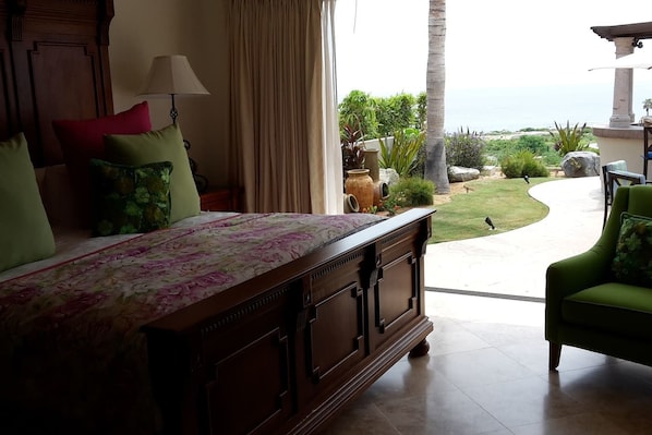 A bedroom with a bed, a nightstand, a lamp, and a view of a garden and ocean.