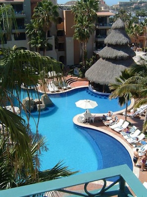 Piscina de resort com espreguiçadeiras e guarda-sóis, cercada por palmeiras e uma construção de colmo.