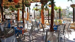 An outdoor restaurant with metal chairs and tables, hanging plants, and a cityscape view. - Great Nautical JR Suites (studio) in Cabo (Cabo San Lucas)