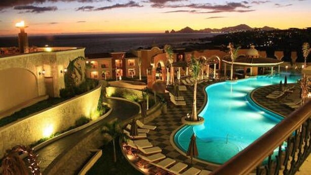 Ein Poolbereich eines Resorts mit geschwungenem Design, umgeben von Liegestühlen und Blick auf den Ozean bei Sonnenuntergang.