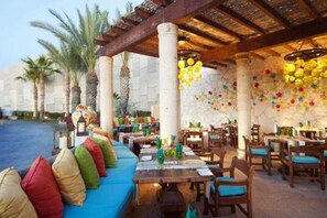 An outdoor dining area with wooden tables and chairs, a blue cushioned bench, and decorative hanging flowers.