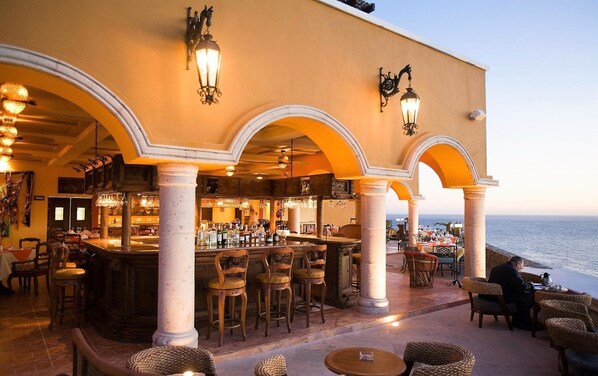 Un área de bar al aire libre con vista al mar, que incluye una barra, asientos e iluminación decorativa.