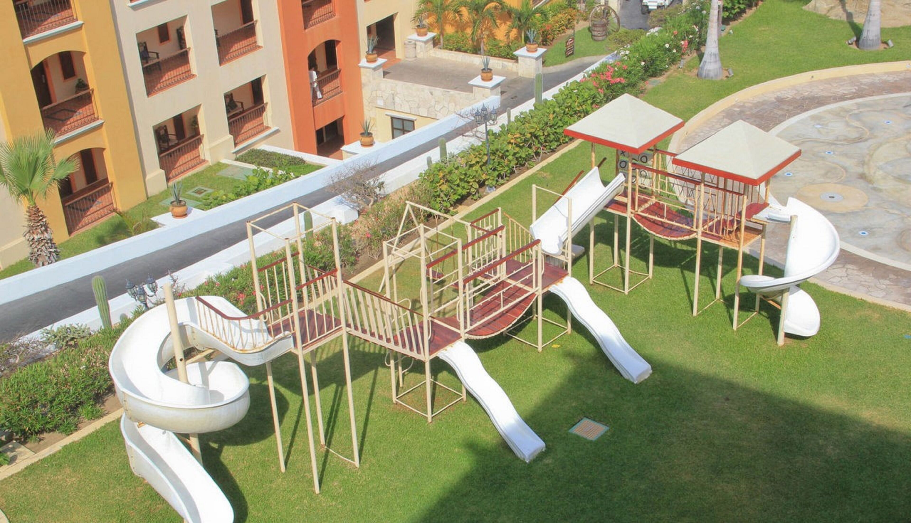 Un parque infantil con toboganes y estructuras para escalar, rodeado de césped cuidado y un edificio con balcones.
