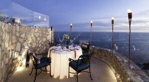 An outdoor dining area with a table set for two, surrounded by stone walls and overlooking a scenic view.