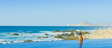 A person standing in a blue infinity pool with a view of the ocean and rocky terrain in the background.