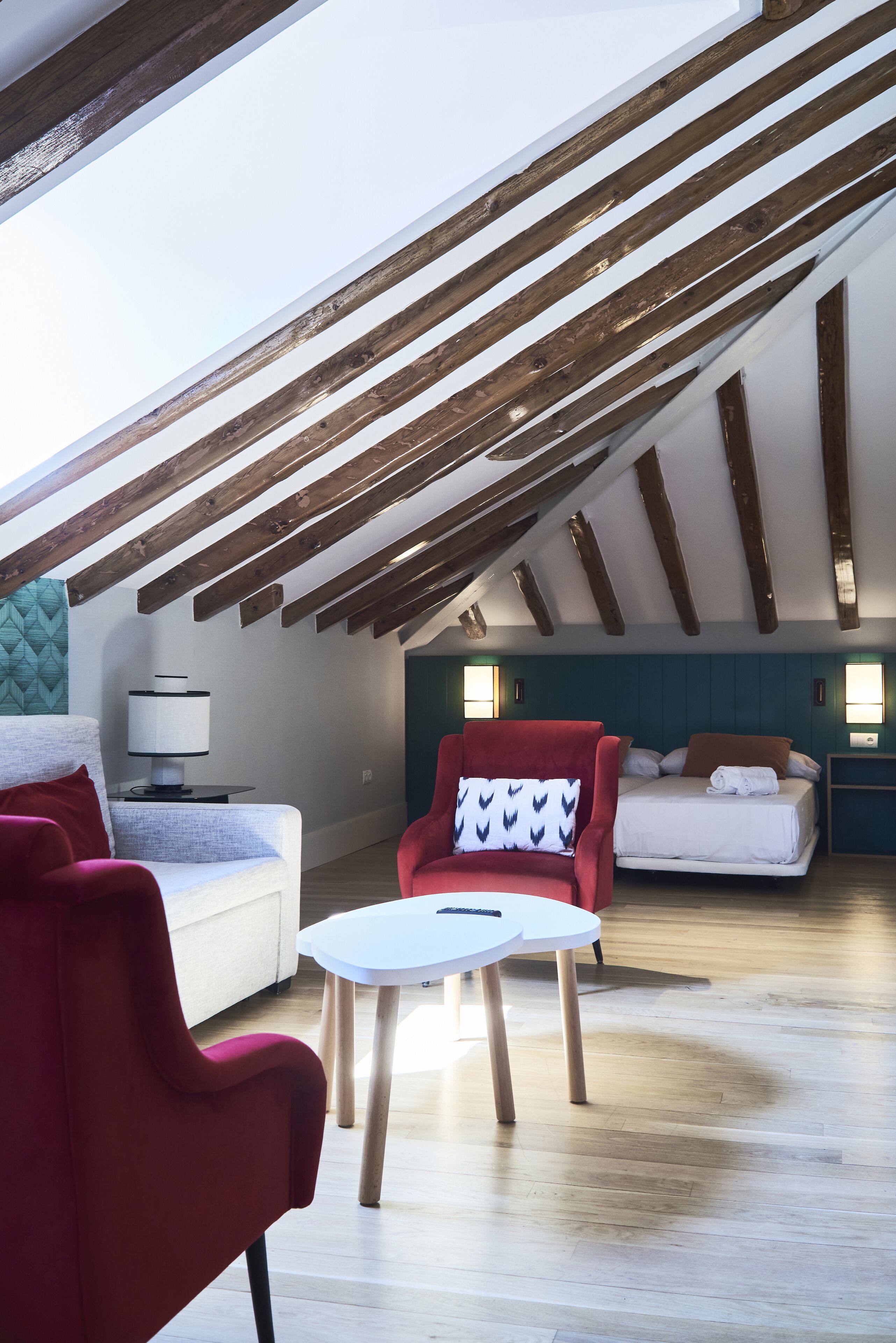 Un amplio dormitorio en el ático con techo inclinado, vigas de madera, una cama, un sillón rojo, una mesita y una lámpara.