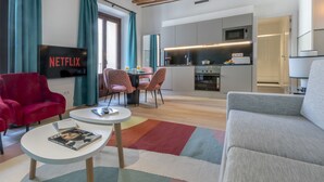 Una sala moderna con un sillón rojo, un sofá gris, un televisor mostrando 'NETFLIX', una cocina al fondo y un tapete colorido.