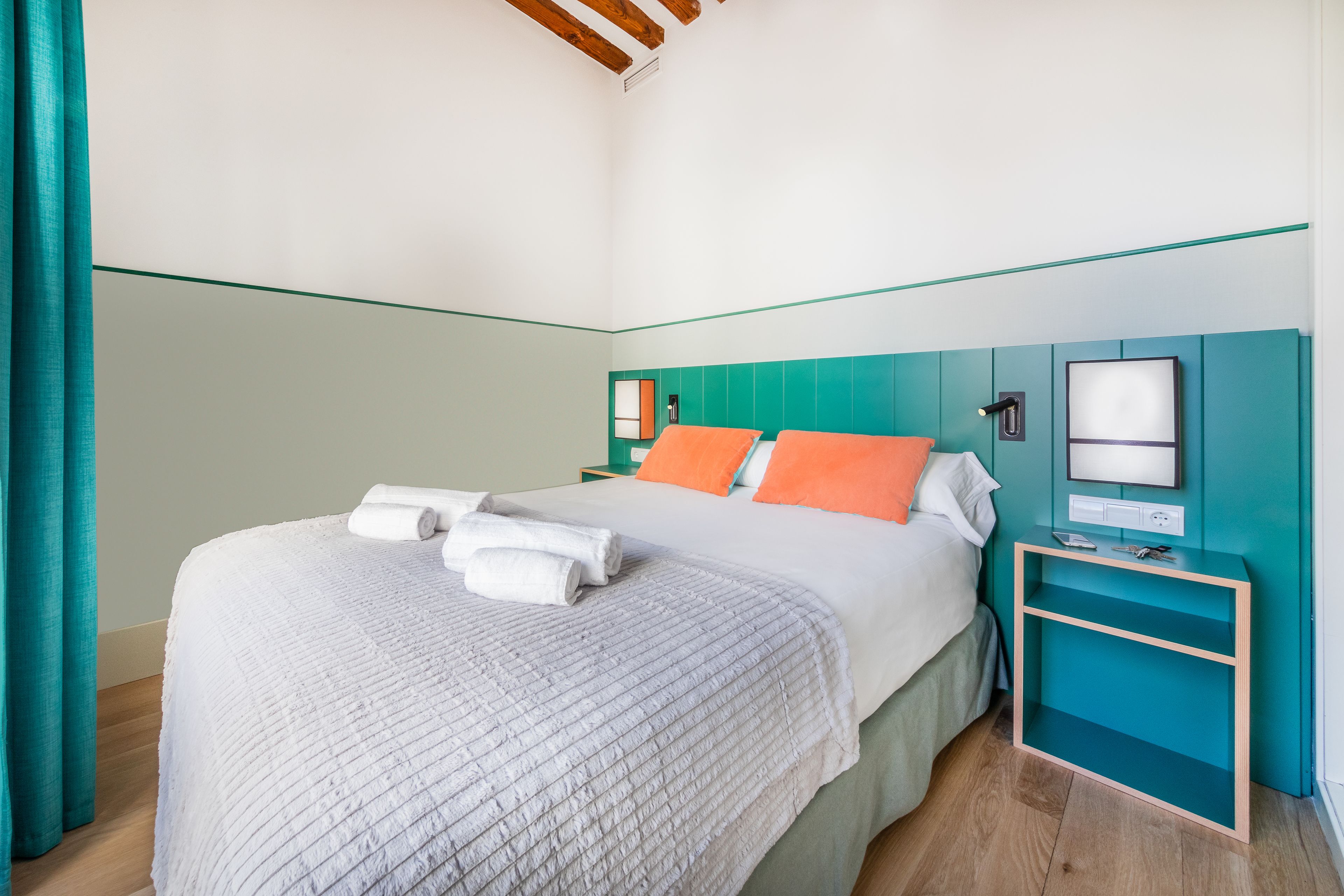 Un lit bien fait, avec une literie blanche et orange, une table de chevet turquoise sur laquelle se trouve un téléviseur à écran plat, et un plancher en bois.