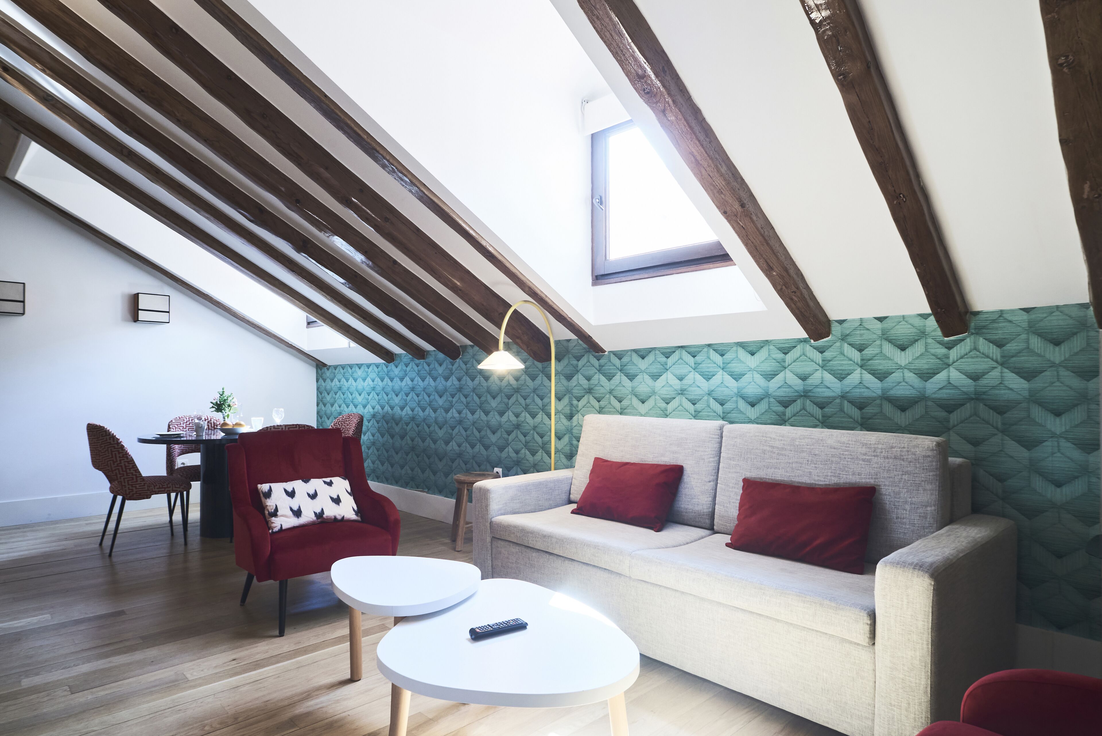 Une chambre moderne de style loft, avec un plafond en pente, des poutres apparentes, un coin repas avec une table et des chaises, un canapé avec des coussins rouges et un mur vert à motifs.