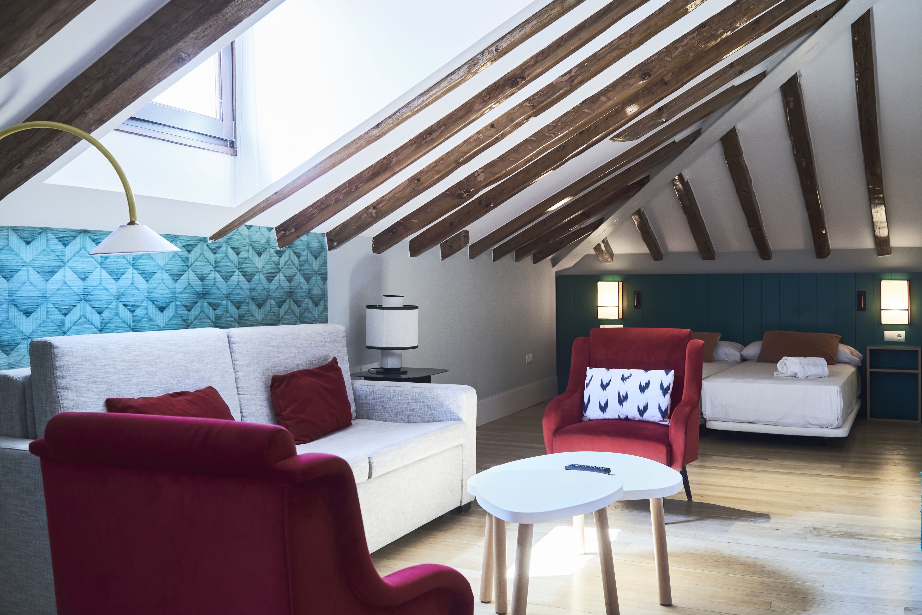 Una habitación estilo loft con techo inclinado, vigas de madera a la vista, un sofá, un sillón rojo, una mesita blanca y una cama con un cabecero azul.