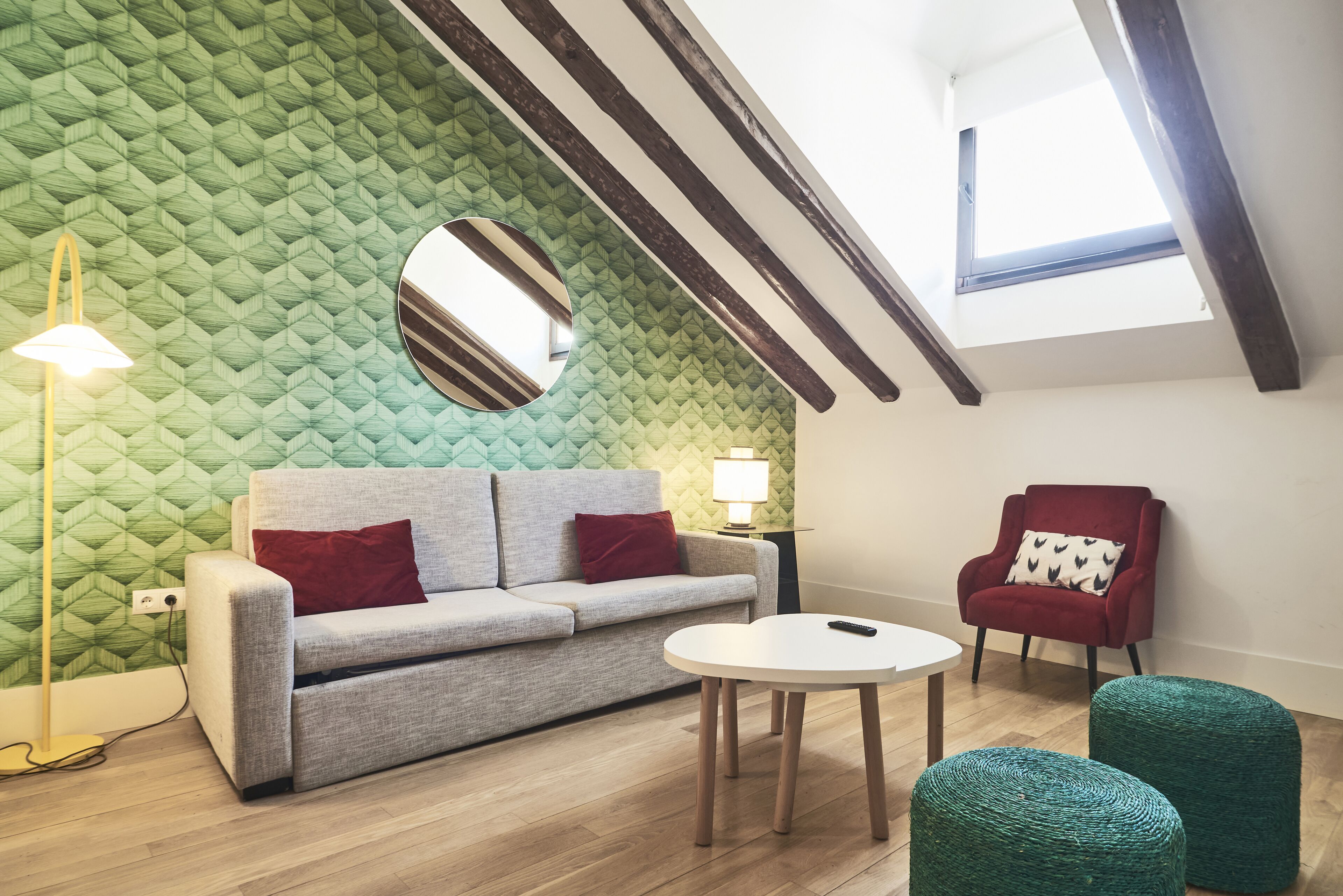 Une pièce avec un mur orné d’un motif vert, un miroir rond, un canapé gris avec des coussins rouges, un fauteuil rouge, une table blanche et une lucarne.