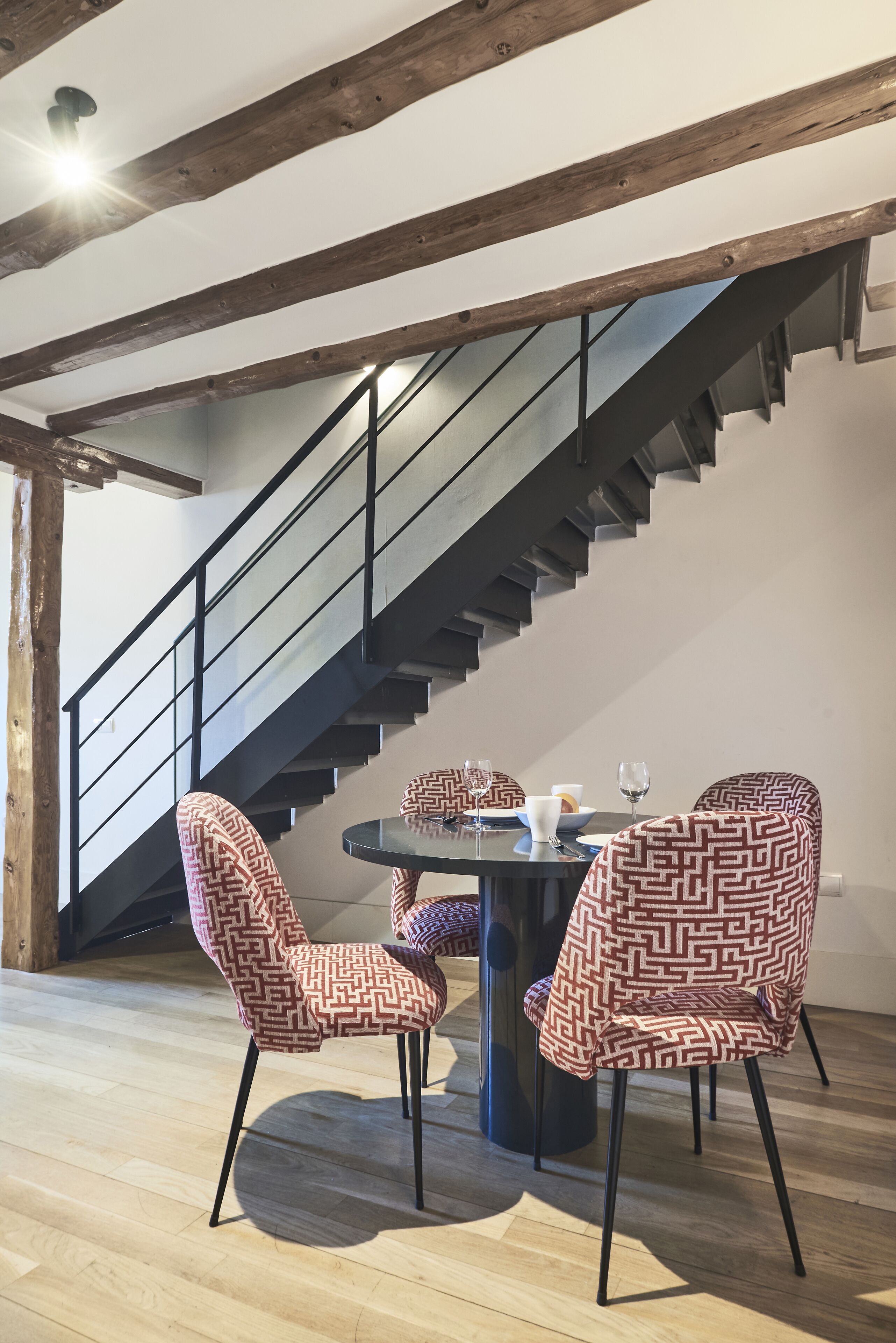 Un área de comedor con una mesa redonda y sillas estampadas, una escalera con barandilla metálica y vigas de madera en el techo.