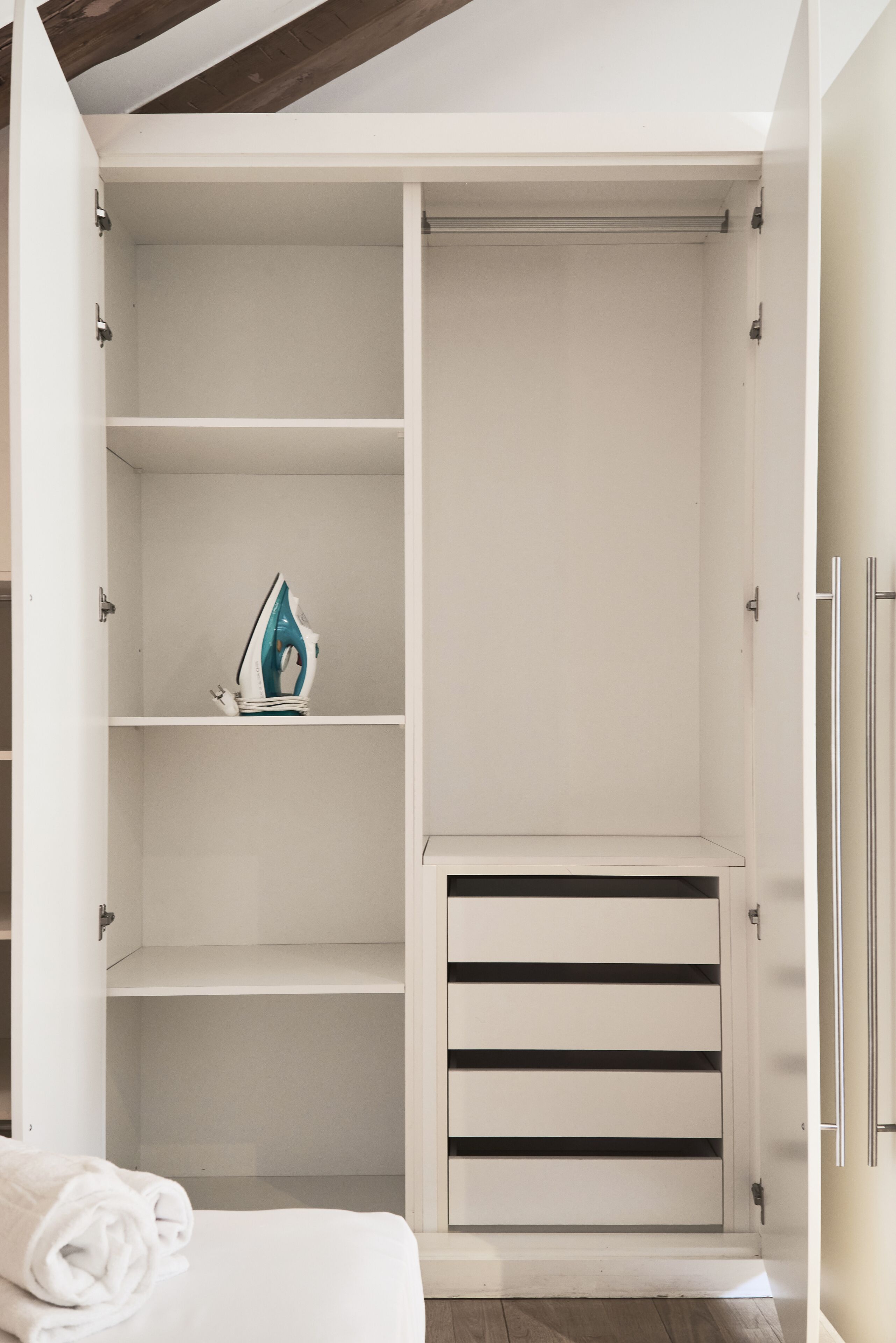 Un placard blanc doté de plusieurs étagères et d’une tringle à vêtements, contenant une serviette blanche pliée et un fer à repasser turquoise et blanc.