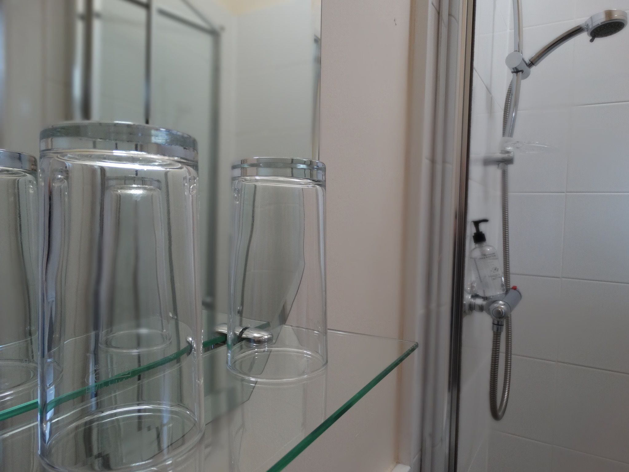 A bathroom with a glass shelf holding clear glass containers, a shower with a handheld showerhead, and a bottle of shampoo.