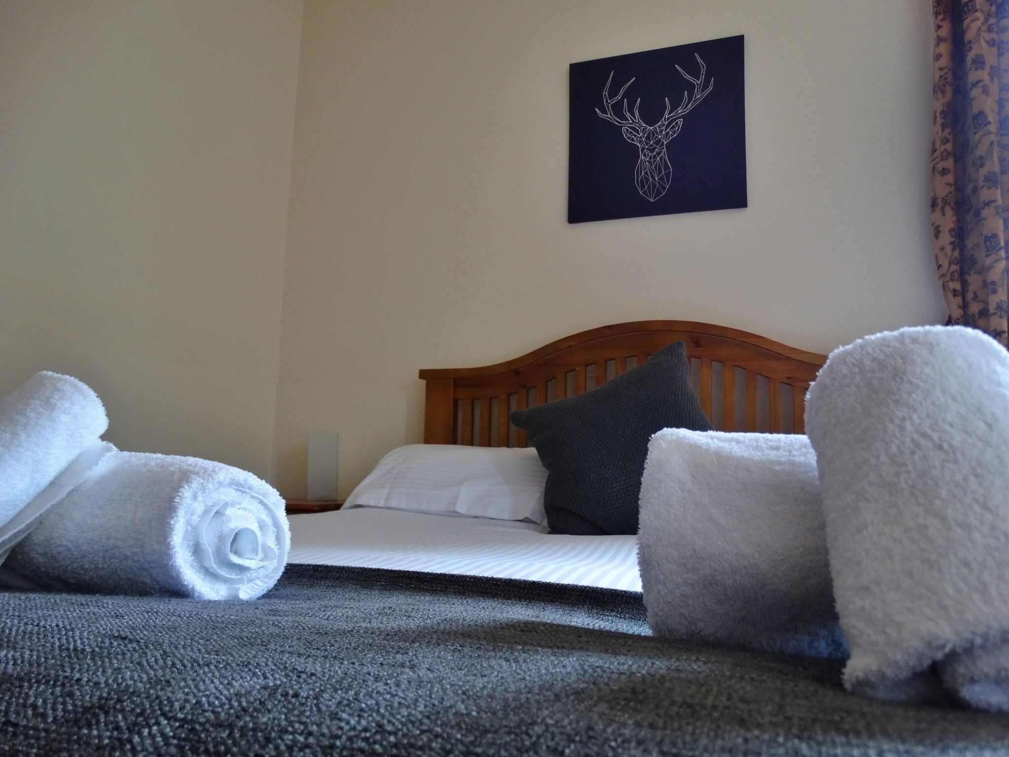 A neatly made bed with white linens, a dark pillow, and rolled white towels on a dark carpet.