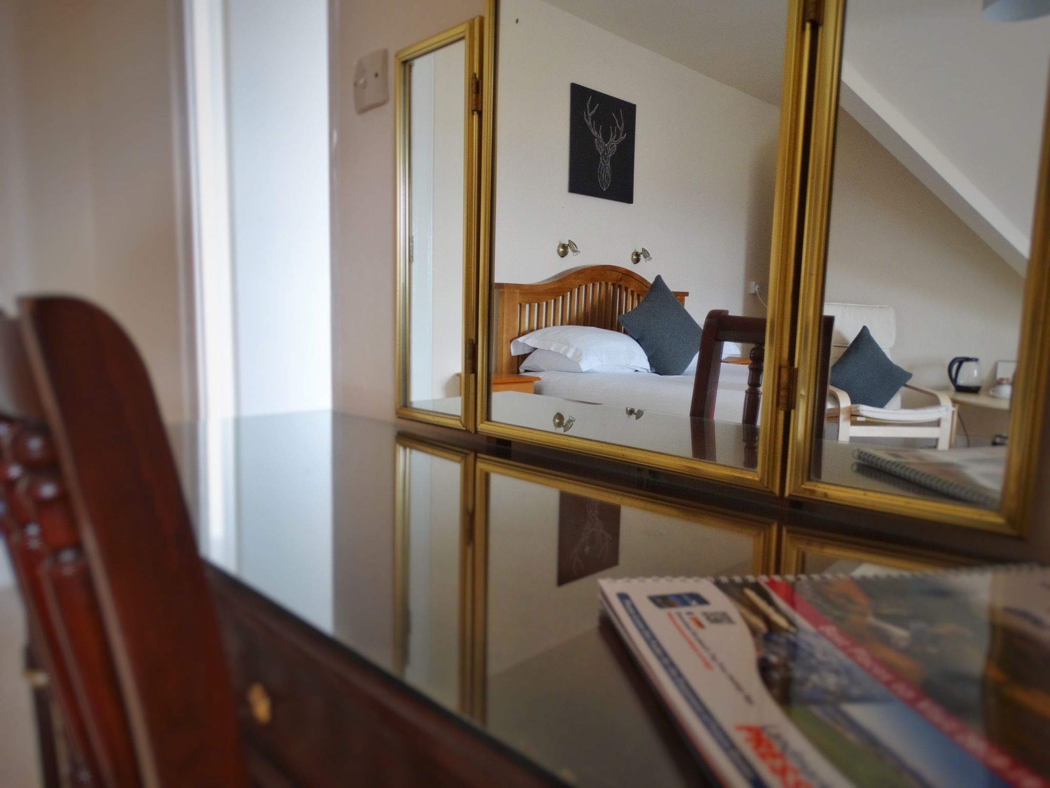 Une chambre d’hôtel avec un grand miroir qui reflète un lit, une tête de lit en bois, un tableau accroché au mur et une petite table sur laquelle se trouvent un livre et une tasse.