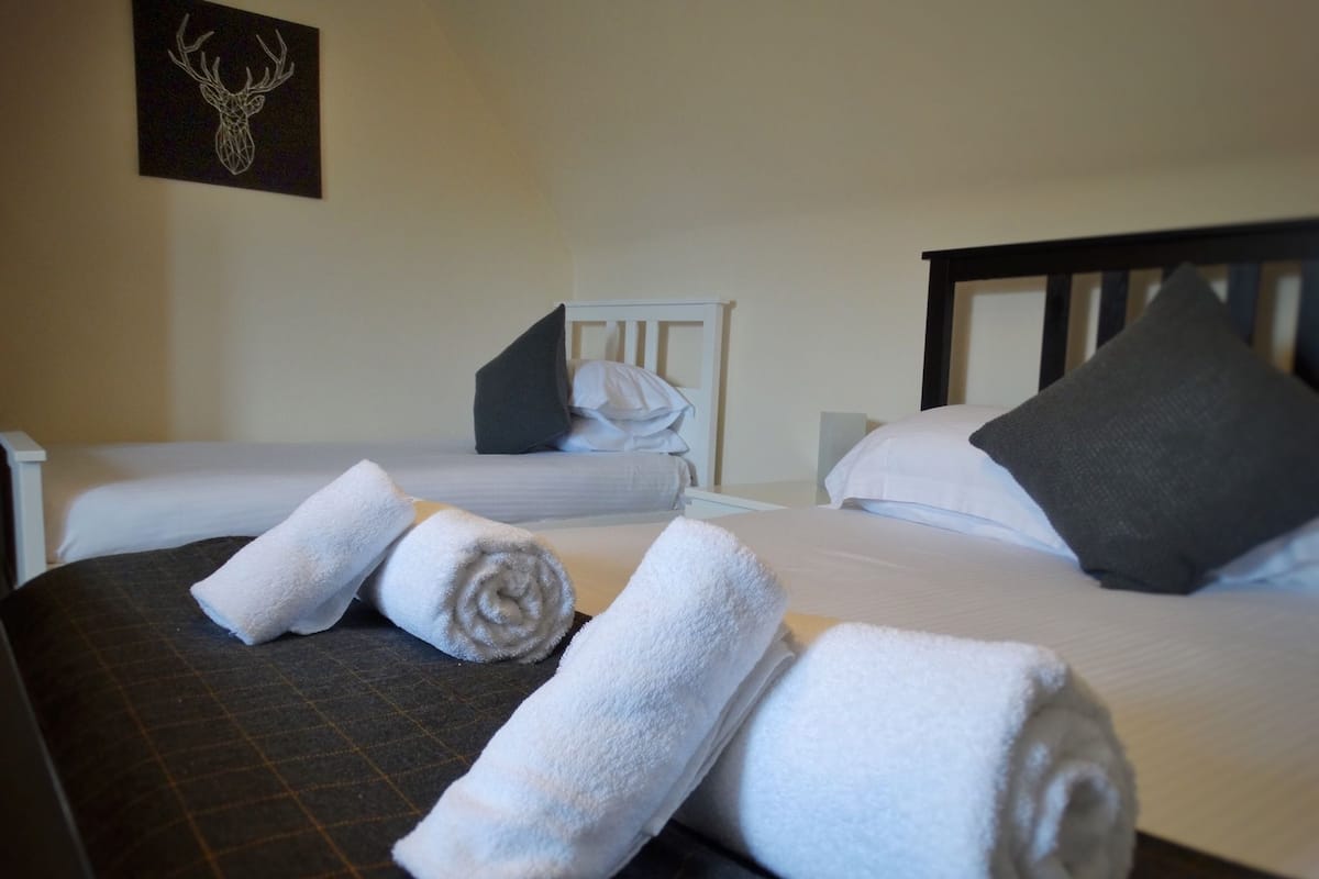A hotel room with two beds, each with white linens and pillows, and a dark patterned bedspread. There are rolled white towels on the beds.