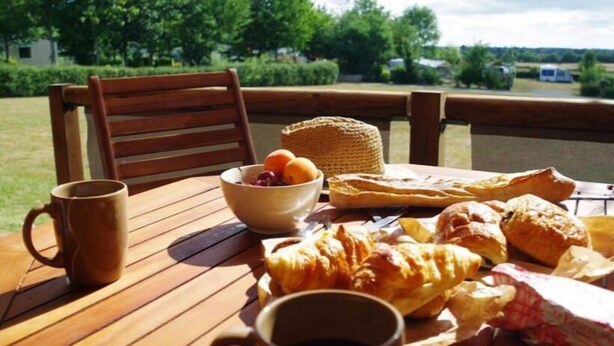Une table en bois, sur laquelle sont disposés un saladier de fruits, des pâtisseries et une tasse, est placée à l’extérieur, offrant une vue sur la verdure et un véhicule au loin.