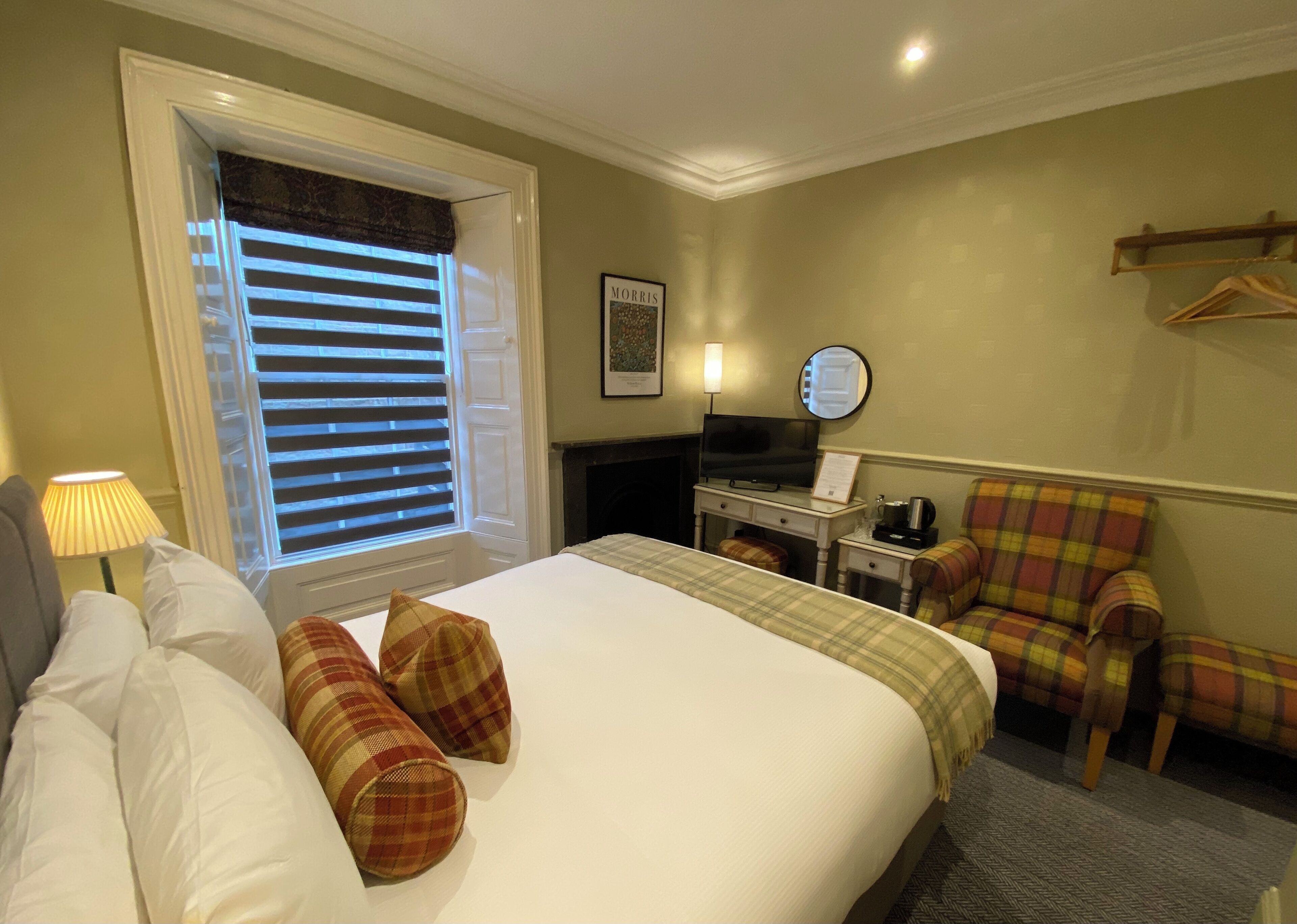 A hotel room with a bed, a desk with a television, a chair, and a window with blinds.