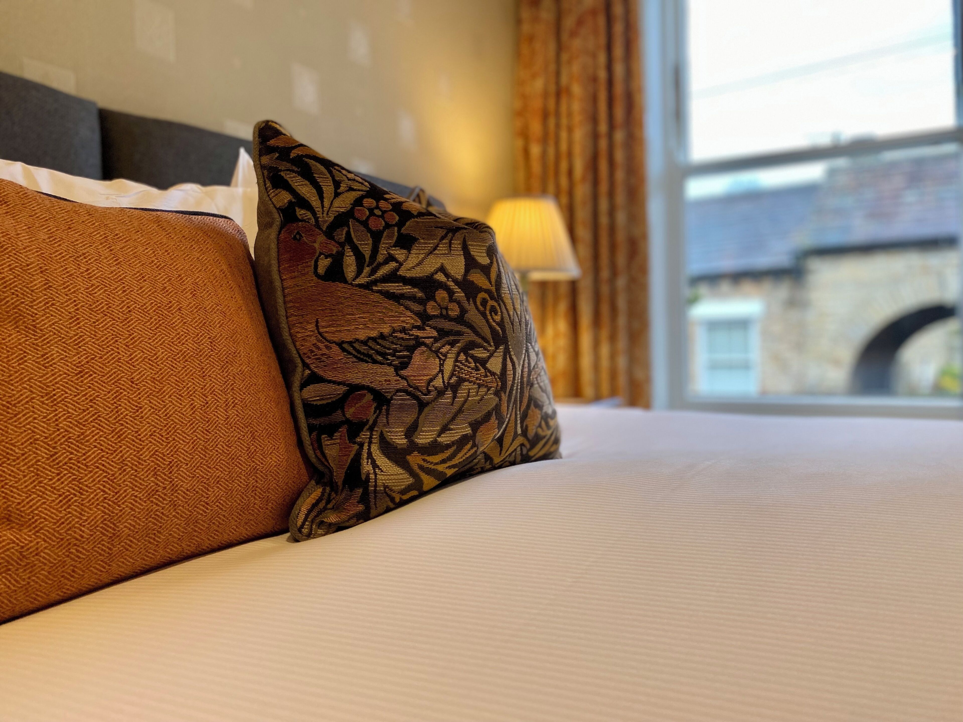 Una cama con dos almohadas, una lisa y otra con estampado floral, en una habitación con ventana y lámpara.