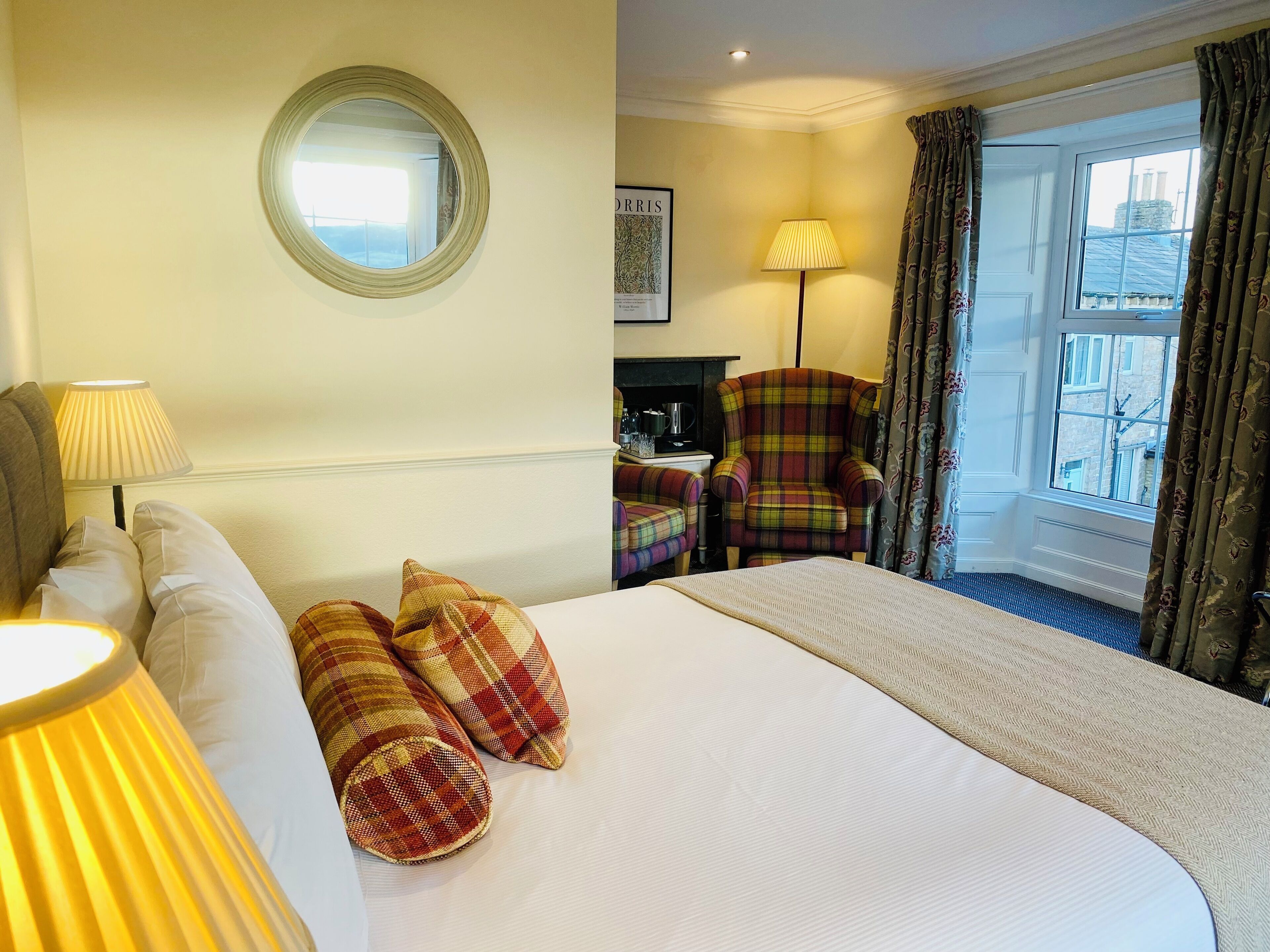 A hotel room with a bed, two lamps, a chair, a desk with a coffee maker, a mirror, and a window with curtains.