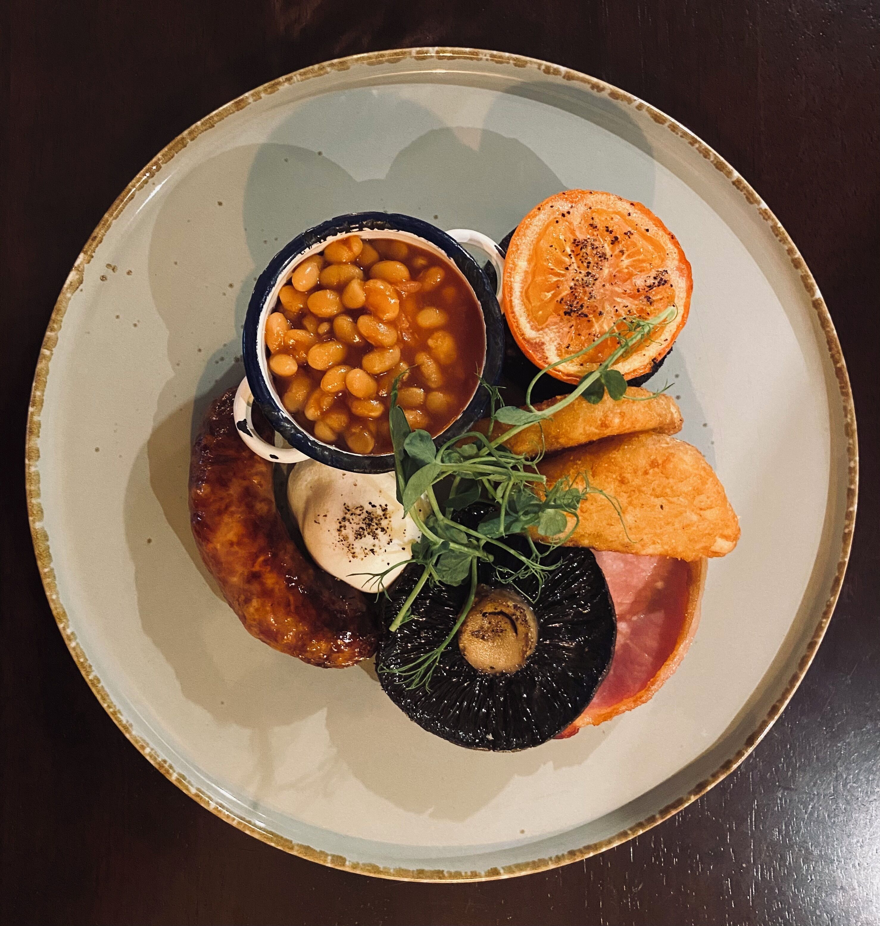 A plate with baked beans, a slice of orange, a mushroom, and a piece of meat.