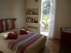 A bedroom with a bed, a bookshelf, a window with curtains, and a small table with water bottles. - Chambre d'hôtes La Villa Du Petit Port (Martigues)