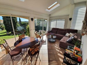 A spacious living area with a large sliding glass door leading to a garden, a brown sofa, a coffee table, and a dining table with chairs. - Chambre d'hôtes La Villa Du Petit Port (Martigues)