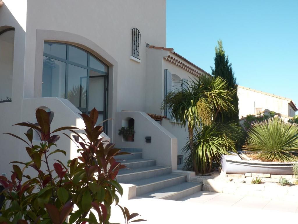 A white building with a staircase leading to a door, surrounded by various plants and a clear sky.
