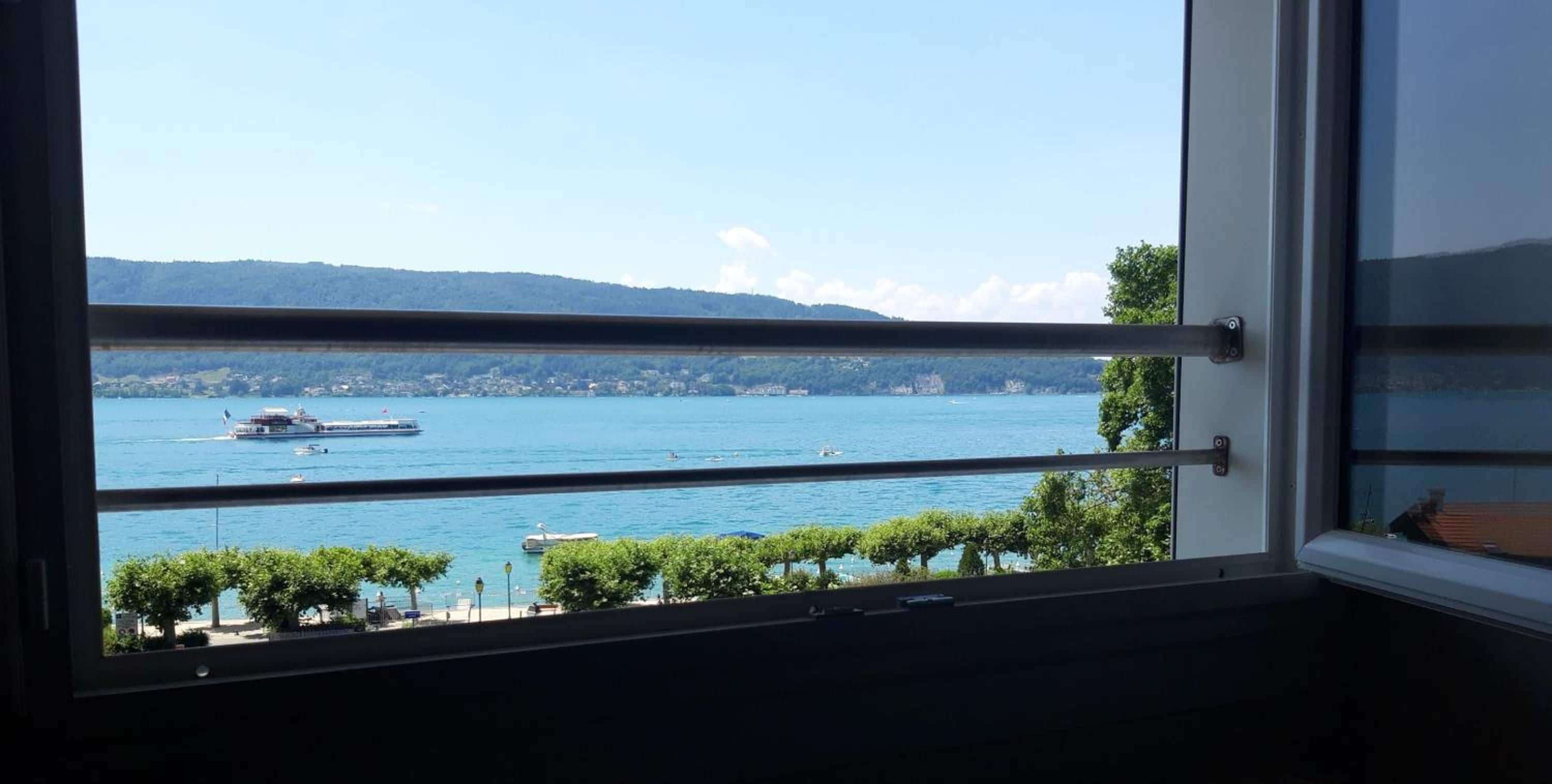 Blick aus einem Fenster auf einen See mit Booten, eine entfernte Uferlinie und Bäume.