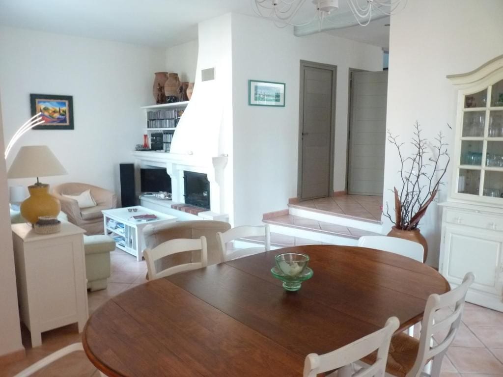 A dining area with a wooden table and white chairs, a small side table with a lamp, and a fireplace in the background.