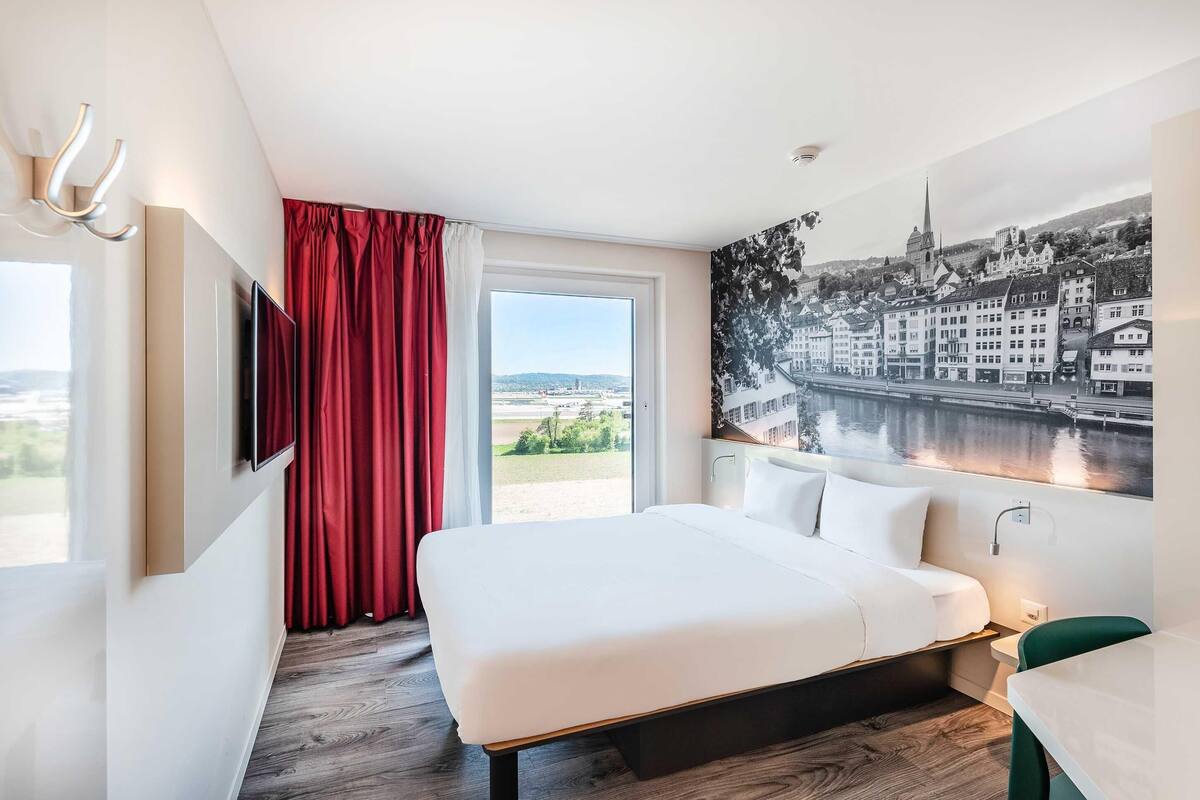 A hotel room with a bed, a TV, a window with a red curtain, and a cityscape painting.