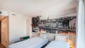 Habitación de hotel con dos camas, un escritorio y vistas a la ciudad a través de una ventana.