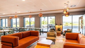 Un moderno hall de hotel con sofás naranjas, mesas redondas y ventanas amplias que dan a un estacionamiento.