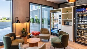Un área de estar moderna con asientos, una mesa de centro, una planta en maceta, una máquina expendedora y un refrigerador lleno de bebidas.