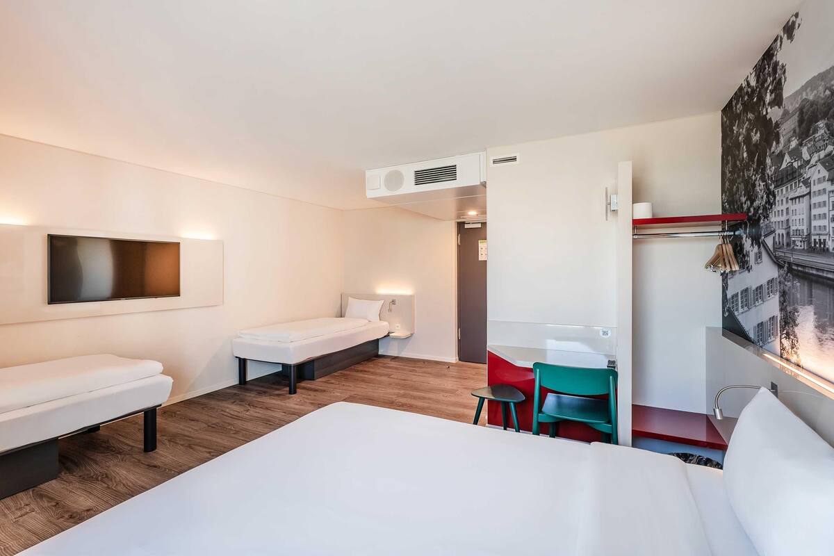 A hotel room with a large bed, a desk, a chair, a small table, a TV, and a wall-mounted air conditioner.