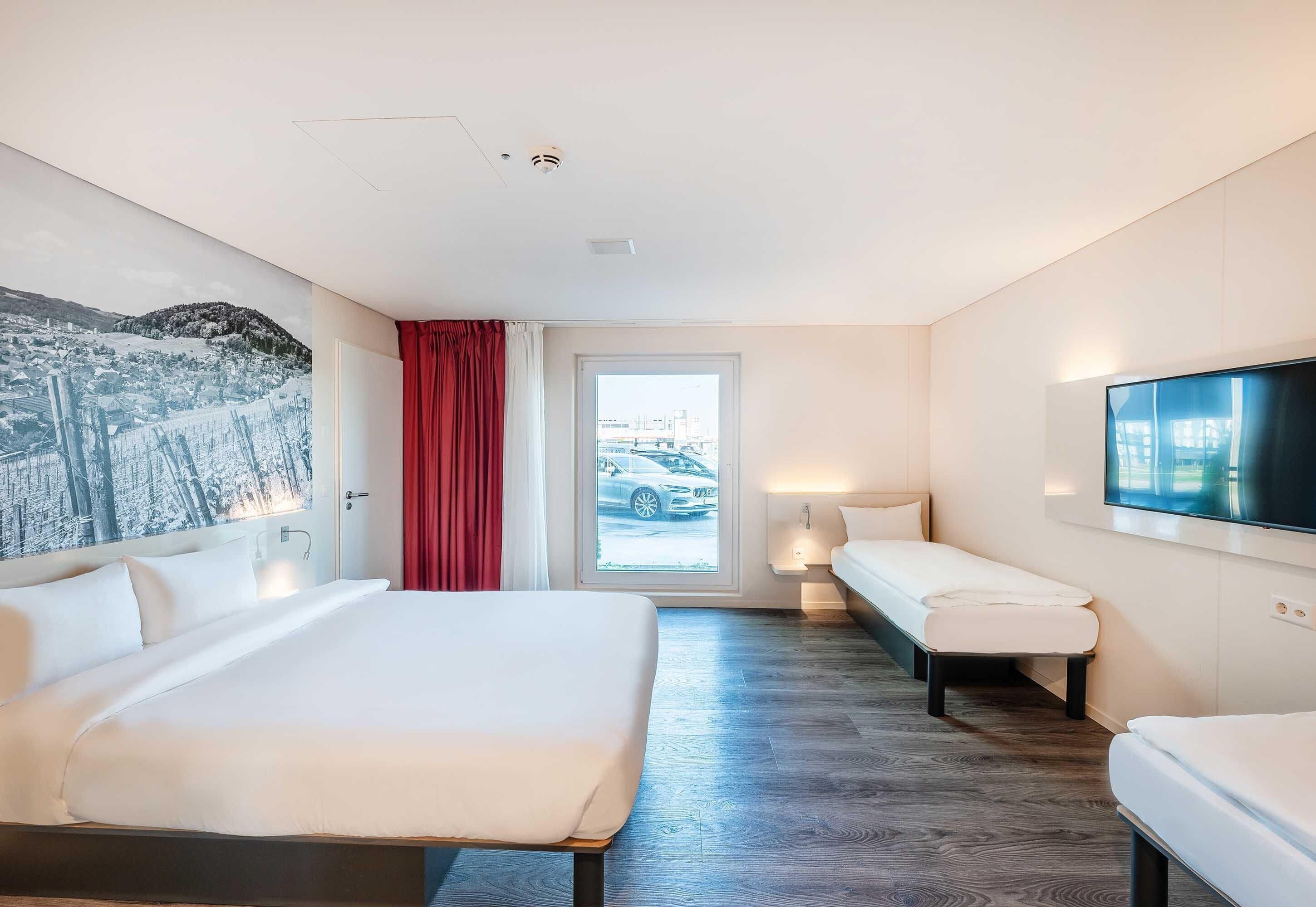 Ein Hotelzimmer mit einem großen Bett, einem kleineren Bett, einem Fernseher, einem Fenster mit Blick auf ein Auto und einem roten Vorhang.