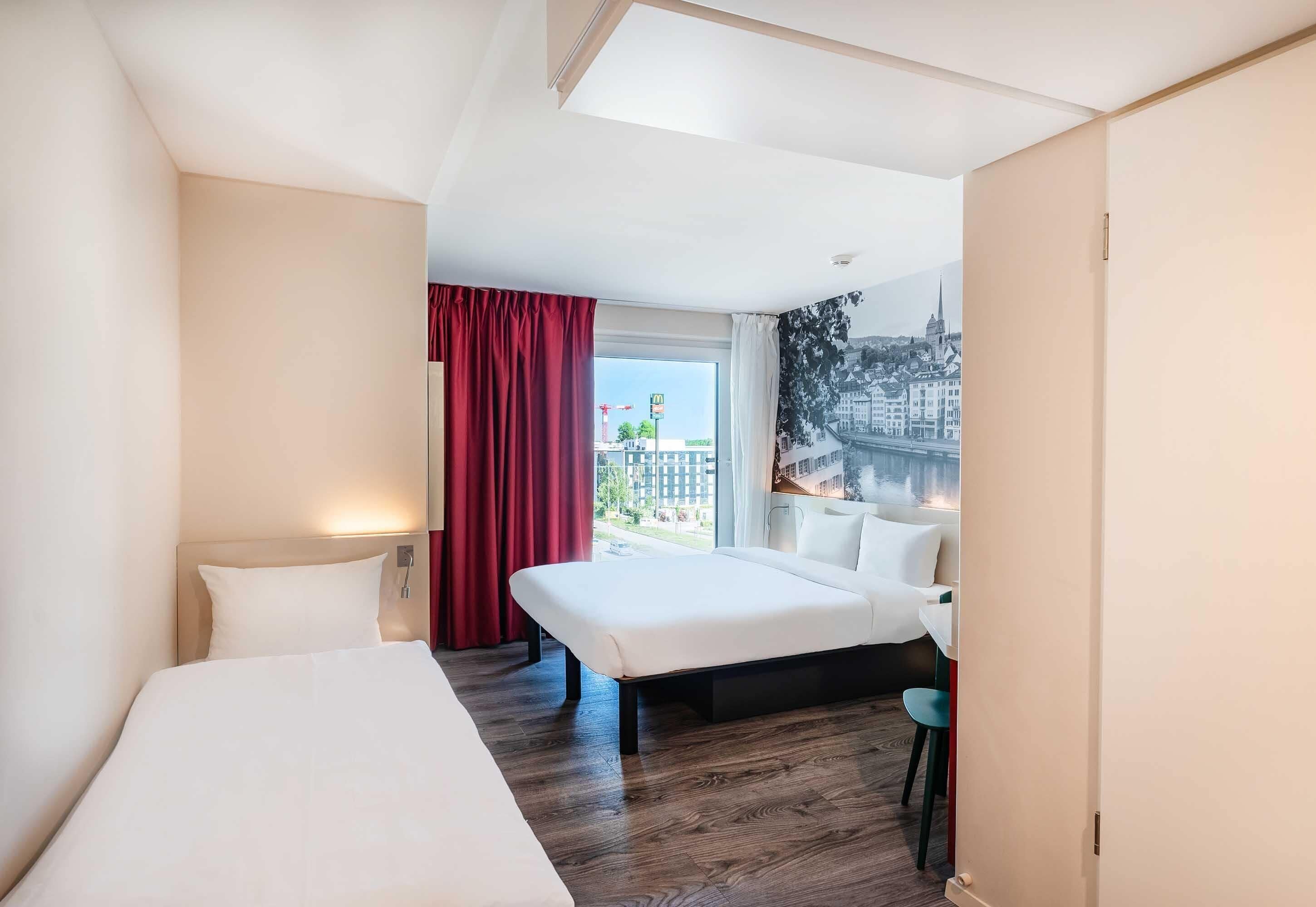 Habitación de hotel con cama, escritorio, silla, ventana y vistas a edificios, además de una cortina roja.