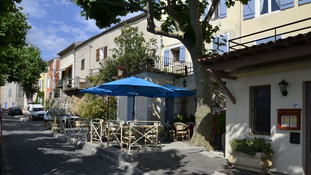 Une rue bordée de voitures garées, avec des terrasses dotées de parasols bleus, et des immeubles avec des balcons et des fenêtres.