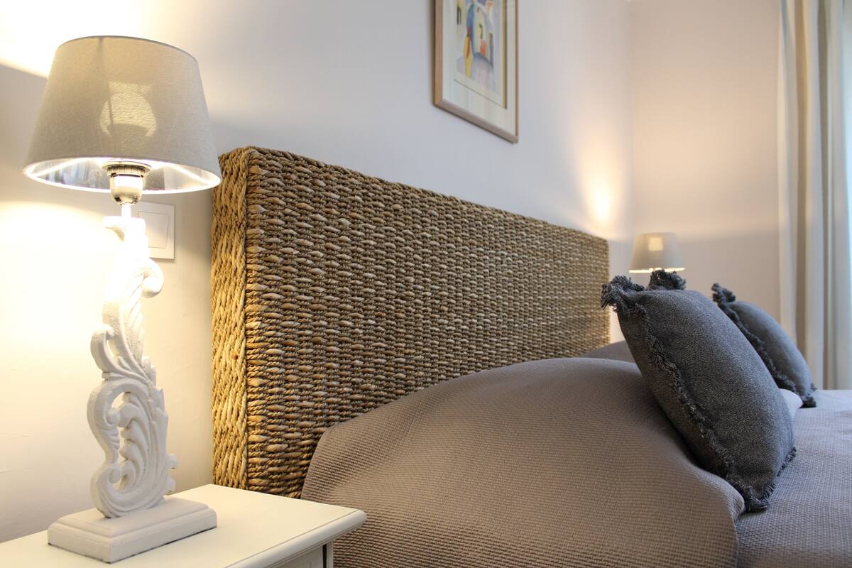 Una cama bien hecha con un cabecero tejido, una mesita de noche blanca con una lámpara y un cuadro en la pared.