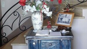 Un escalier orné d’une rampe en métal décorative, un vase rempli de fleurs rouges et un petit tableau encadré posé sur une commode bleue.