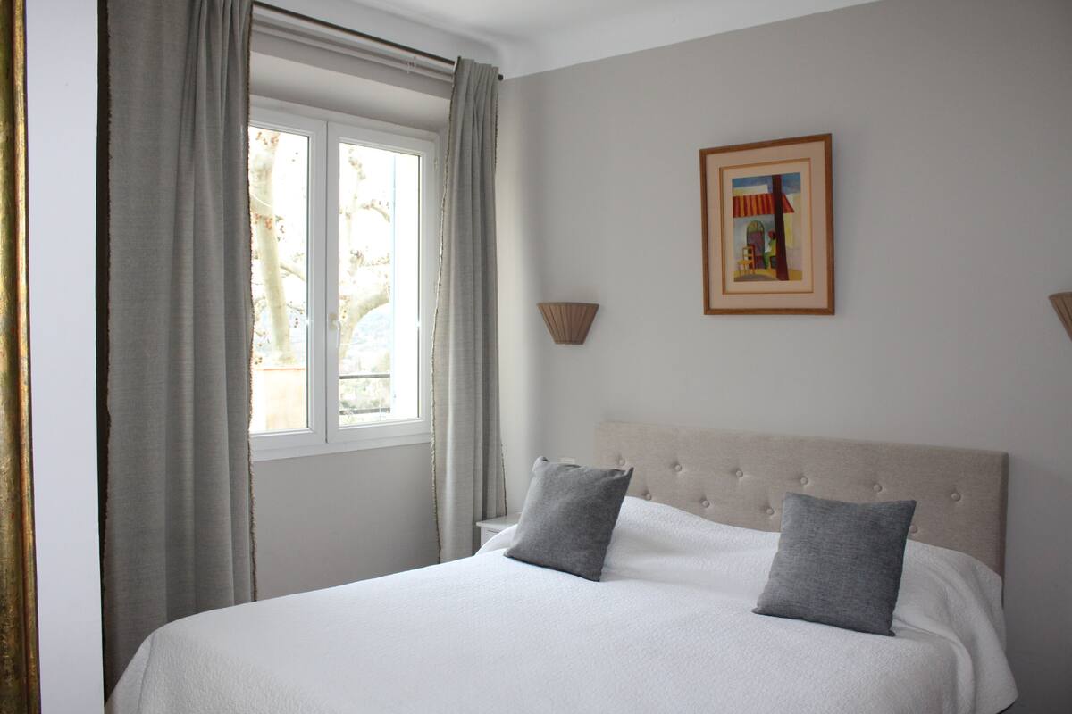 Una cama bien hecha con cabecera, una ventana con cortinas y un cuadro en la pared.