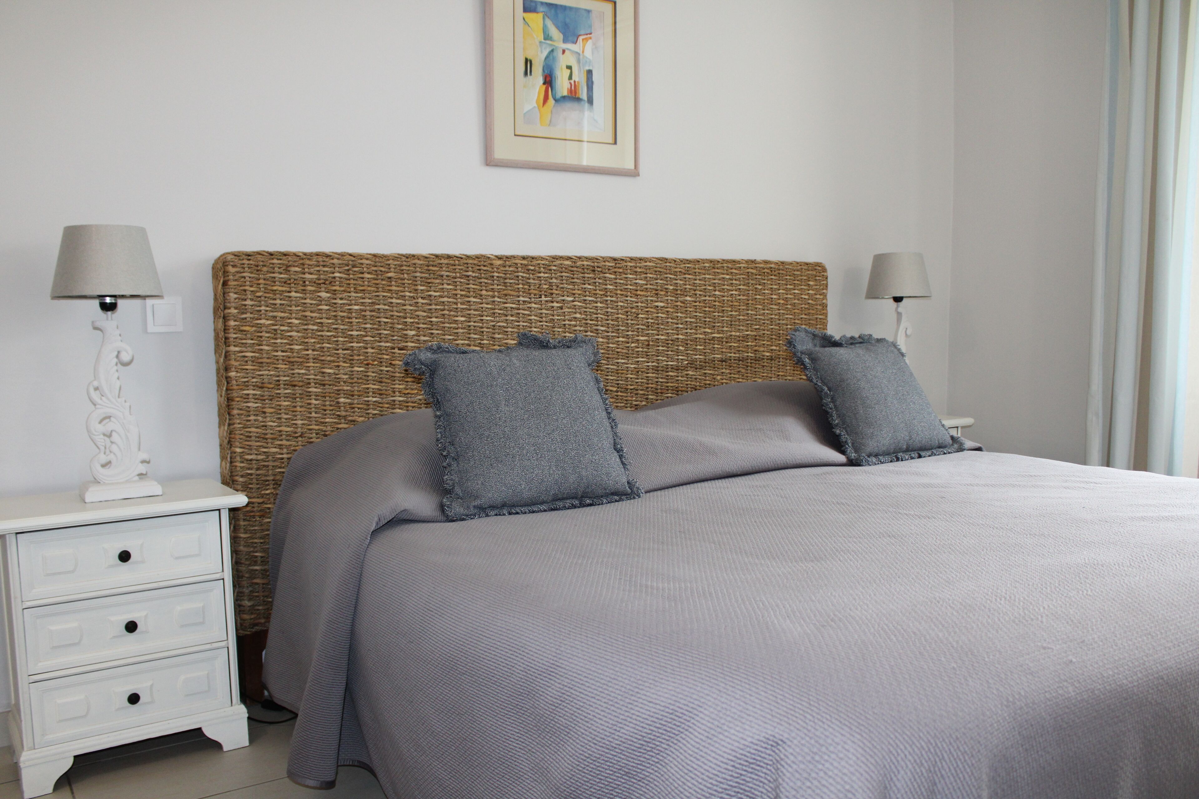 Una cama bien hecha con colcha gris y almohadas, un cabecero de mimbre y dos mesitas de noche con lámparas.