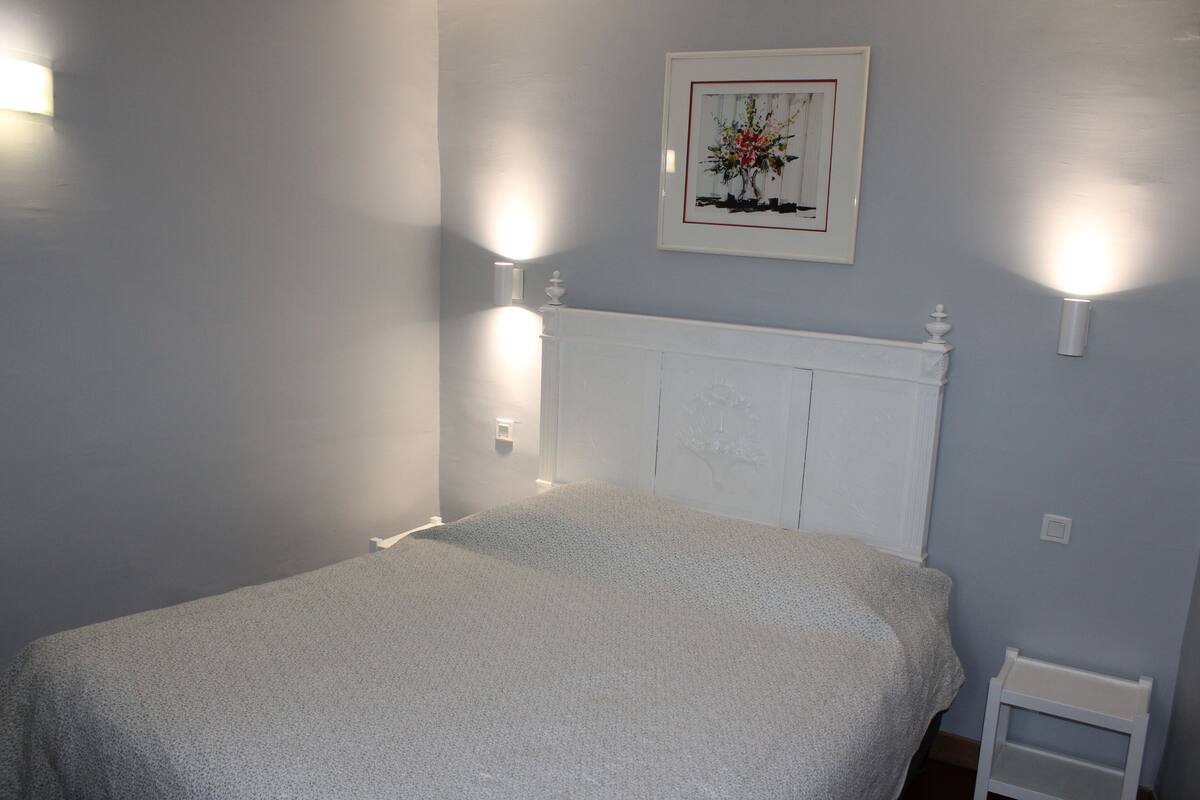 Un dormitorio con una cama blanca, una mesita de noche blanca y un cuadro en la pared.