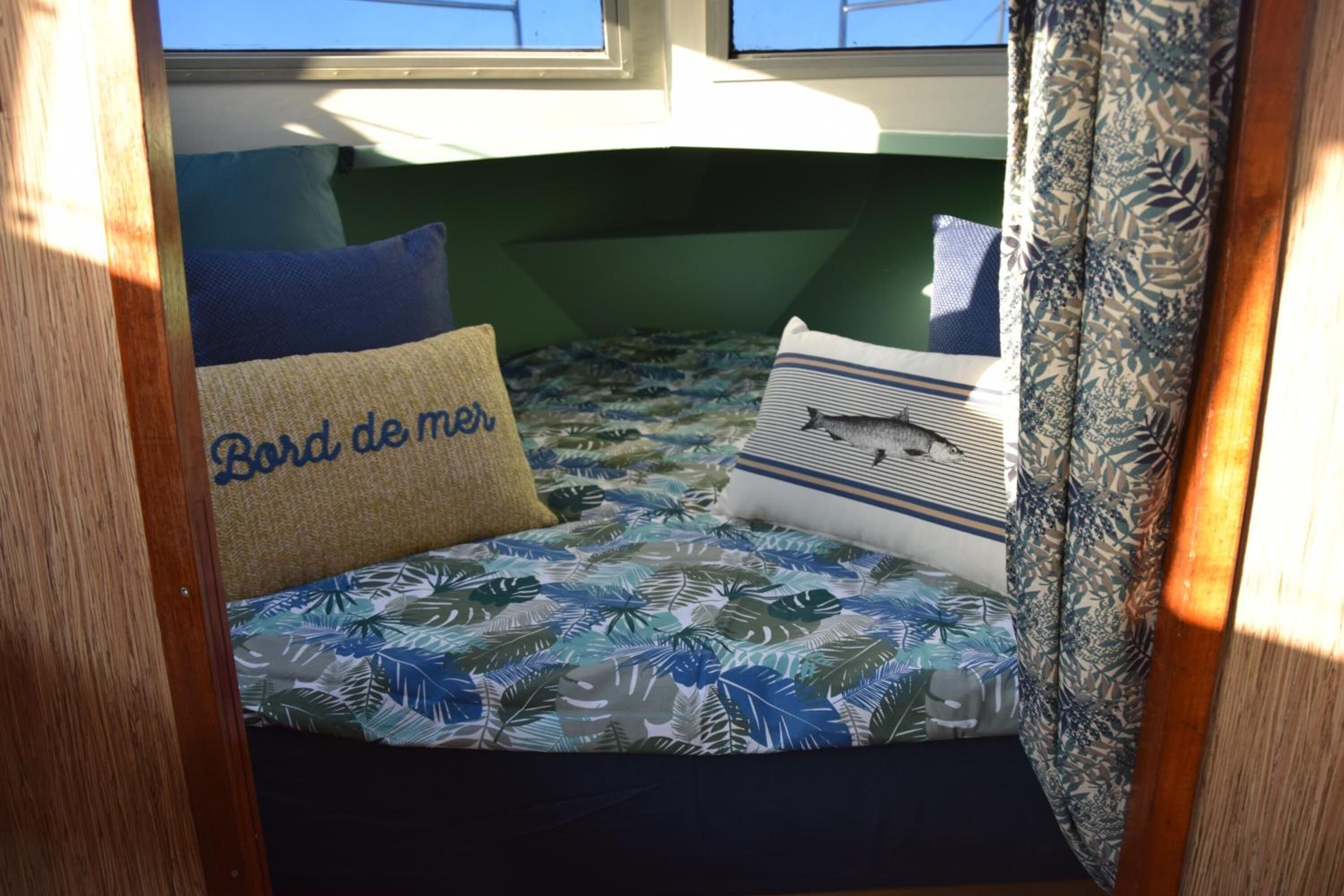 Een kleine, gezellige kamer met een bed, voorzien van beddengoed met een tropisch bladmotief, een kussen met de tekst 'Bord de mer' en een gestreept kussen.