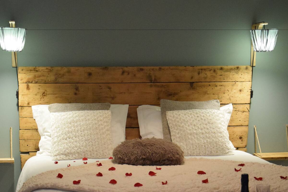 Una cama con cabecera de madera, ropa de cama blanca, un edredón blanco esponjoso y pétalos de rosa esparcidos sobre la cama. Hay dos copas de vino y una botella en la mesita de noche junto a la cama.