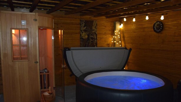 A wooden sauna room with a large, dark-colored hot tub filled with water, a wooden door, and a wall clock. - BAIE EN BULLES - Les Portes du Hable (Cayeux-sur-Mer)